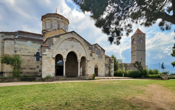 Trabzon Ayasofya Müzesi’nin dış cephesi ve çan kulesi; Komnenos dönemine ait Bizans mimarisi, geniş narteks kemerleri ve kubbeli ana mekân ile birlikte açık bir bahçe alanı.
